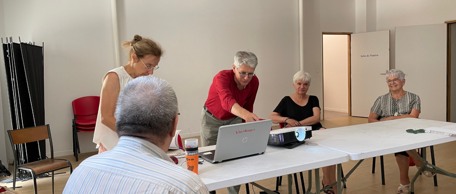 Un petit groupe de personnes seniors échange autour d’une table, avec un ordinateur portable et un vidéoprojecteur, dans une salle claire et accueillante.