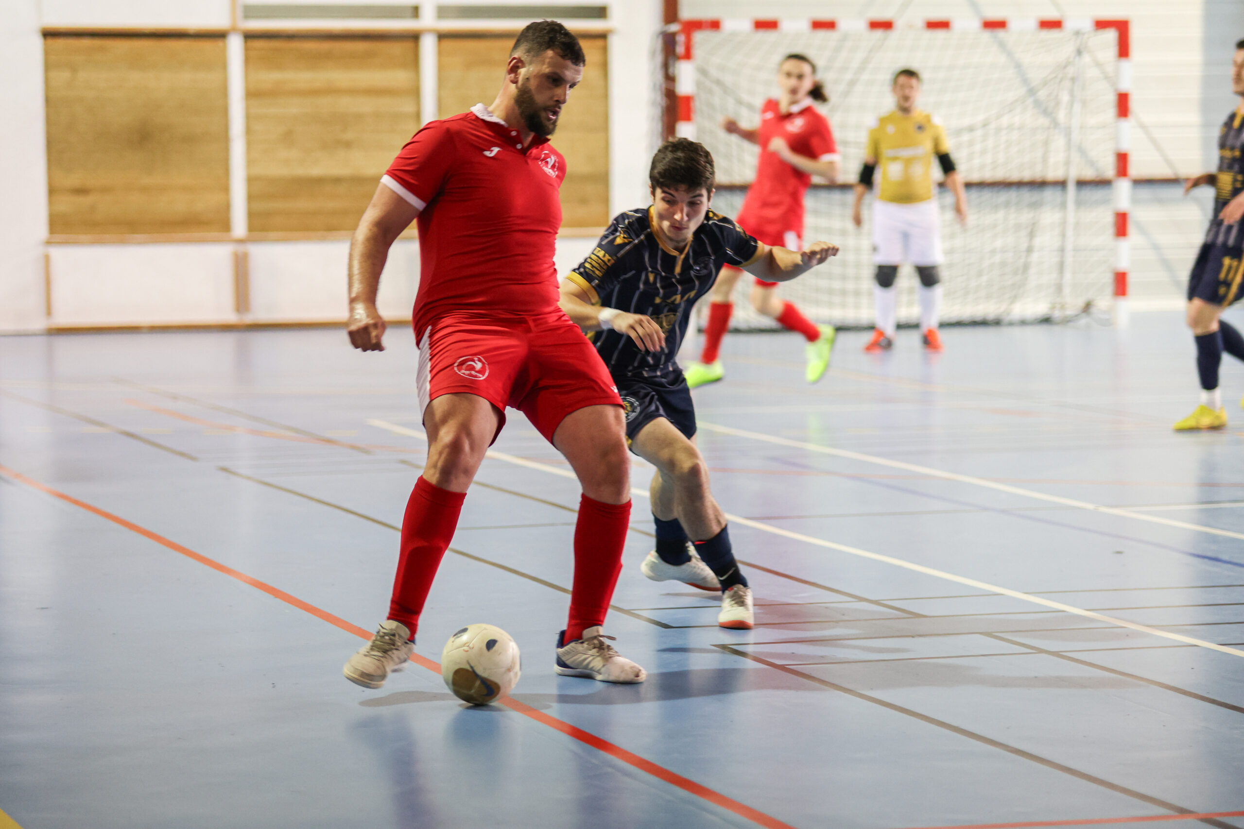 Un joueur en tenue rouge contrôle le ballon sous pression d’un adversaire en tenue bleue, sur un terrain de futsal en salle. D'autres joueurs et un gardien sont visibles en arrière-plan.