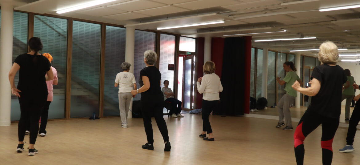 Un groupe de personnes, majoritairement des femmes, participent à un cours de danse dans une salle de sport aux murs vitrés. L’instructrice est en face, guidant les mouvements.