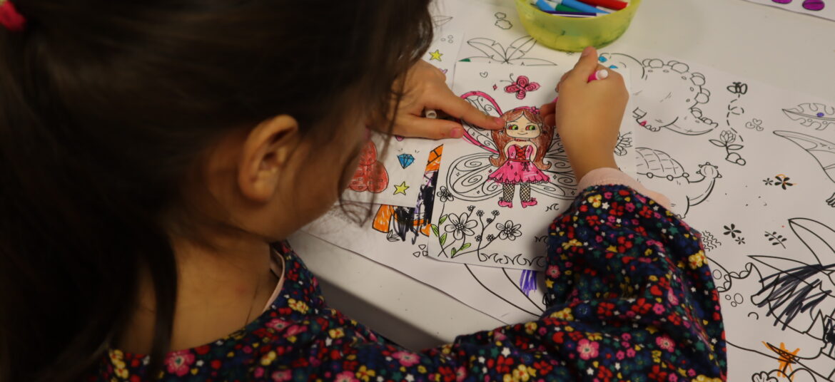 Une jeune fille colorie un dessin de fée sur une feuille, entourée d'autres dessins et de feutres colorés posés sur une table.