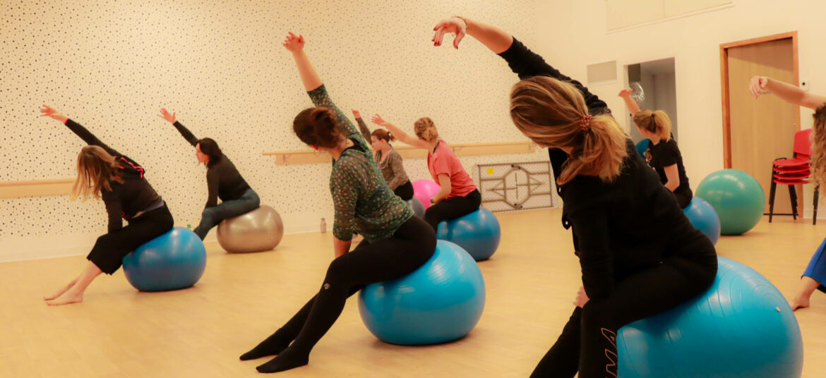 Des femmes participent à un cours de Fit Ball en salle, assises sur des ballons de gym, effectuant un étirement latéral du bras dans une salle claire au sol en bois.