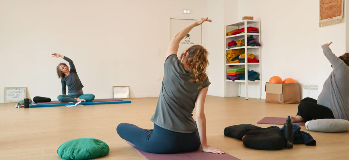 Trois femmes pratiquent une séance de yoga dans une salle lumineuse, guidées par une instructrice assise au fond de la pièce. Toutes effectuent un étirement latéral en position assise.