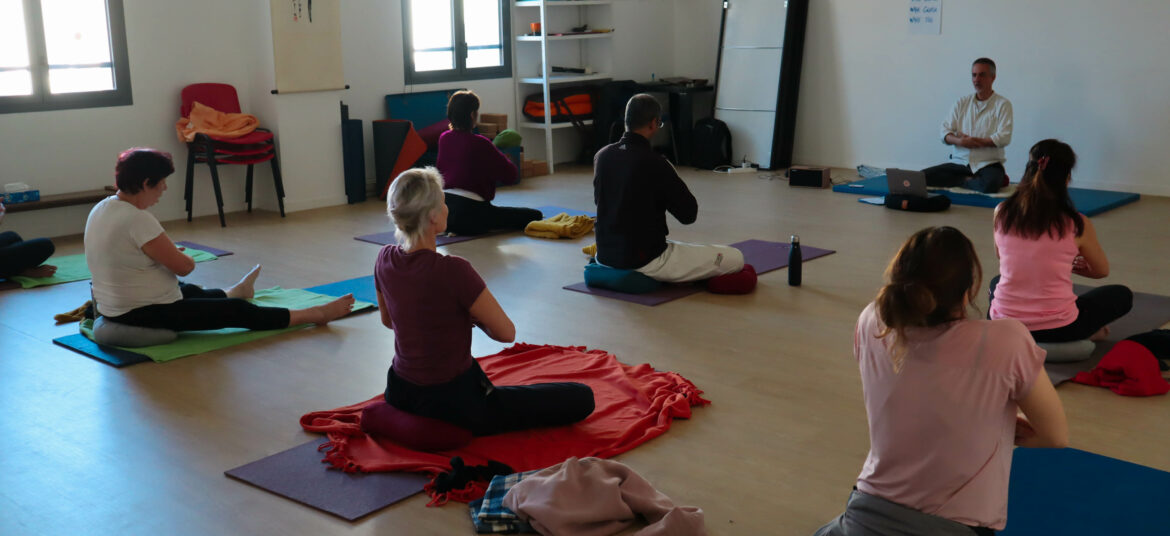 Un groupe de personnes assises en tailleur sur des tapis de yoga, en train de suivre une séance de méditation guidée dans une salle lumineuse. Un enseignant est assis face au groupe, à côté d’un ordinateur portable.