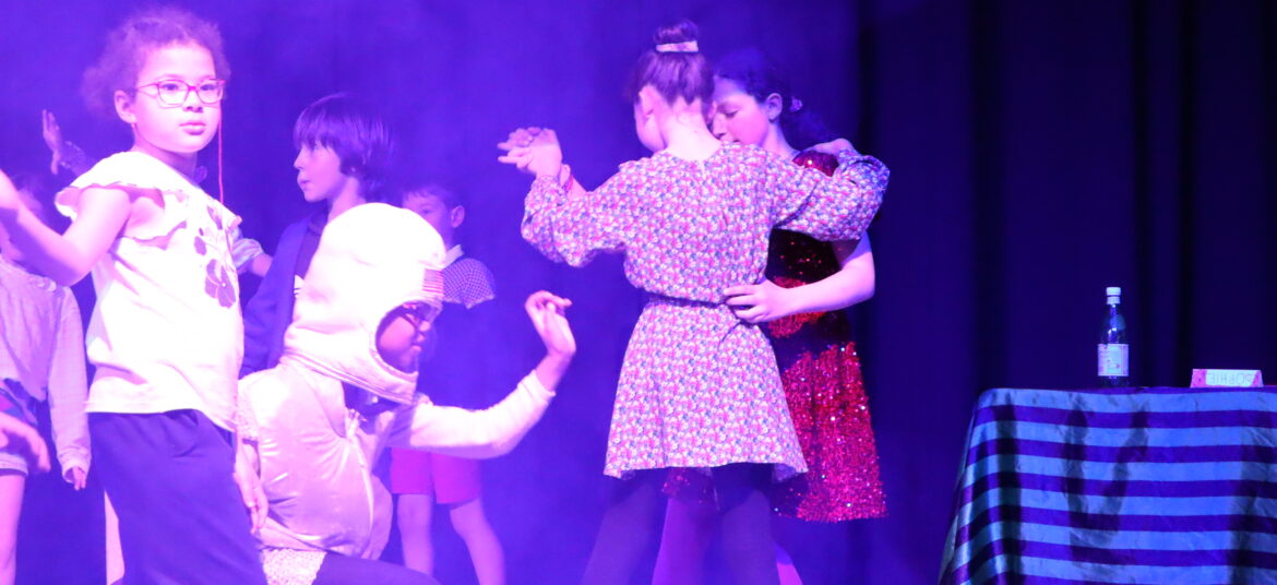 Des enfants en costume jouent une scène de théâtre sur scène, sous des lumières colorées, dans une ambiance créative et expressive.