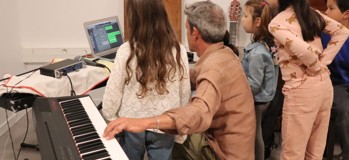 Un adulte et un groupe d’enfants sont rassemblés autour d’un ordinateur et d’un clavier numérique, en train de composer ou d’enregistrer de la musique.