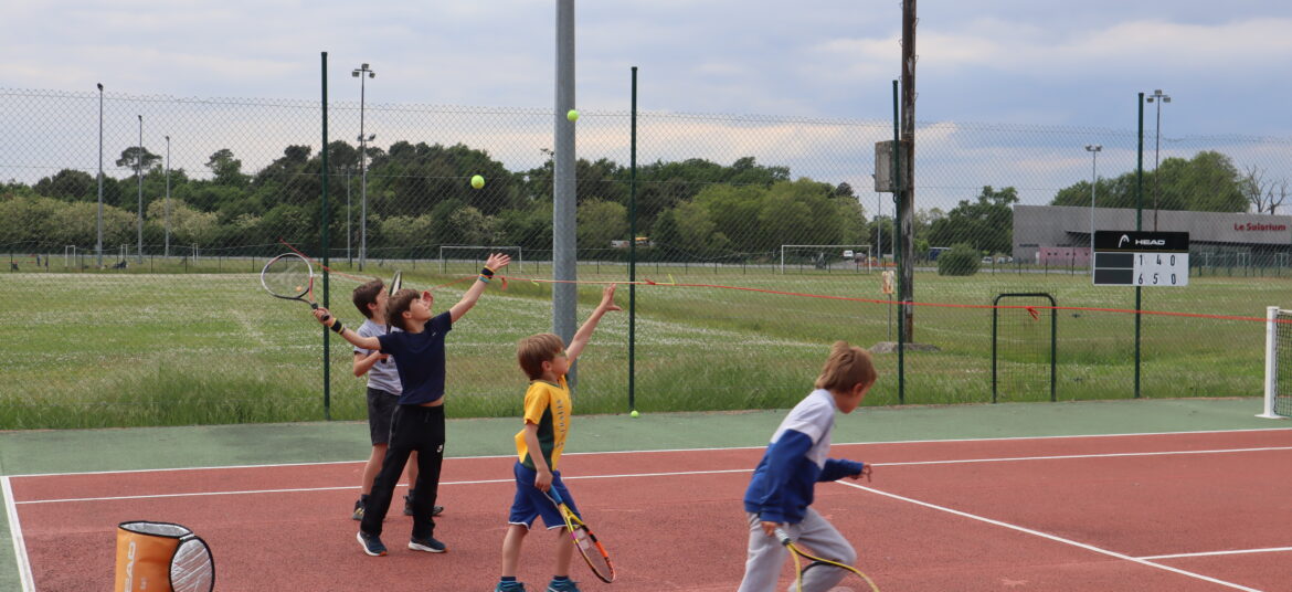 Des enfants en tenue de sport s’exercent au service sur un court de tennis en plein air.