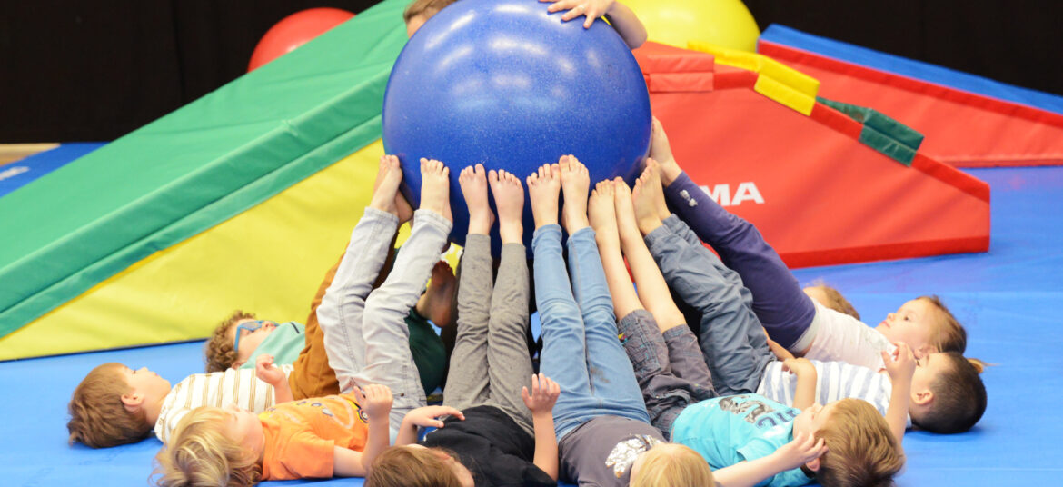 Un groupe de jeunes enfants allongés sur un tapis de gym, les jambes levées pour tenir une grande balle bleue avec leurs pieds, lors d’un atelier de motricité.