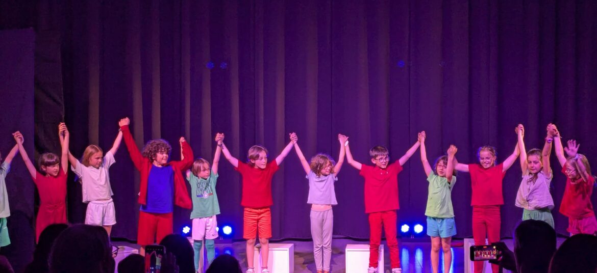 Groupe d’enfants sur scène se tenant par la main lors du salut final d’un spectacle de théâtre aux Coqs Rouges, sous les applaudissements du public.