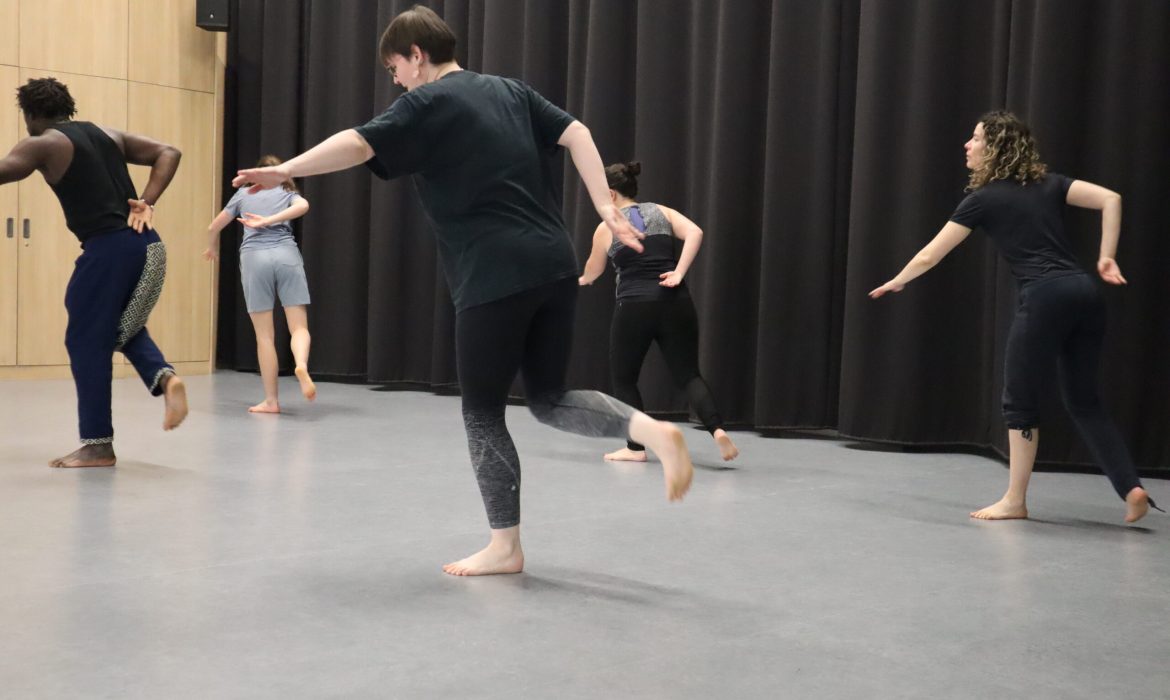 Un groupe d’adultes pieds nus en pleine session de danse africaine, dans une salle de danse avec sol gris et rideaux noirs.